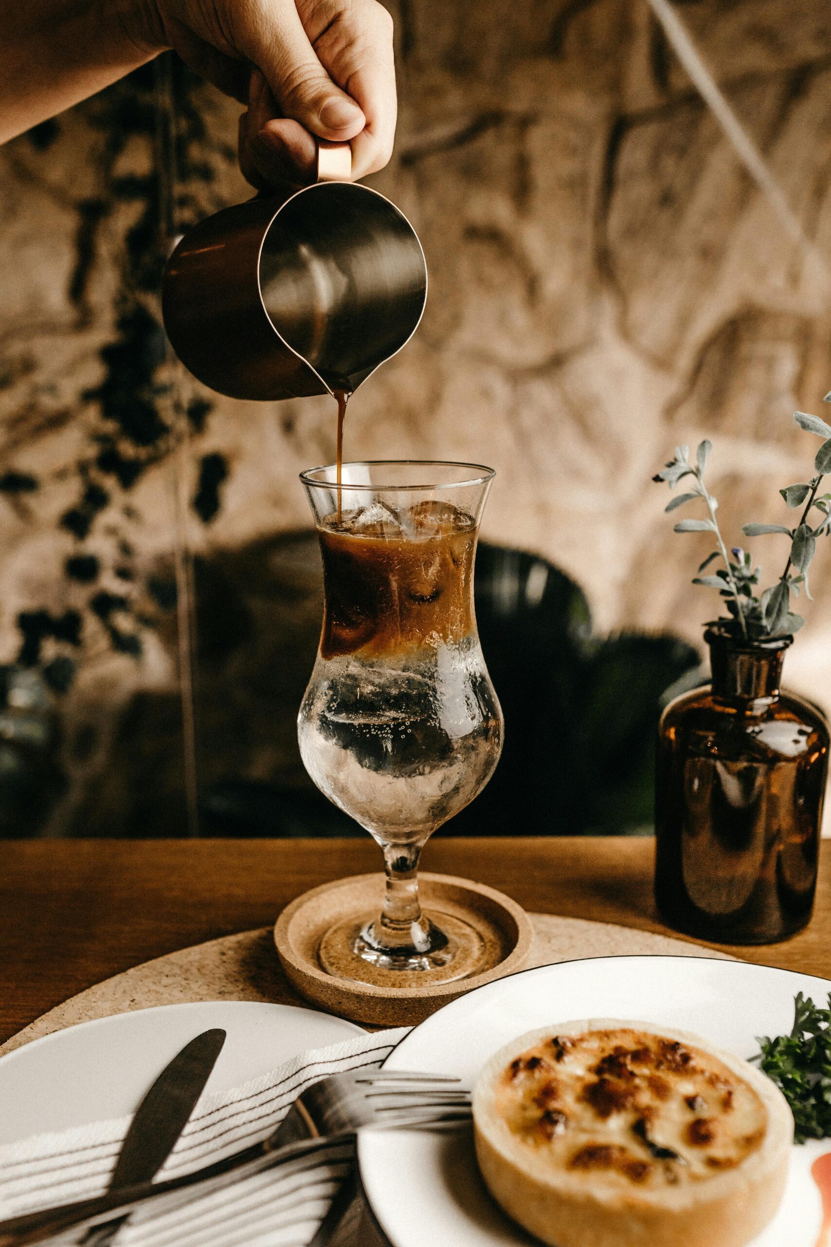 Hand pouring iced coffee into a glass on a table with a pie and cutlery in a cozy cafe.